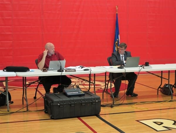 Two people sit behind a table. 