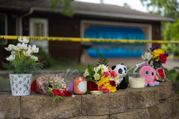 A memorial with flowers, candles and stuffed animals lines a retaining wall outside a house.