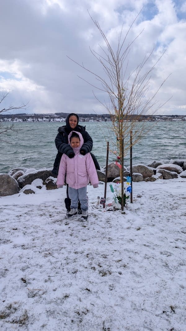A woman and her young daughter stand next to a tree growing on a lake shoreline. Snow has blanketed the ground.