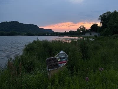 A boat sits in high grass near a lake as the sun goes down.