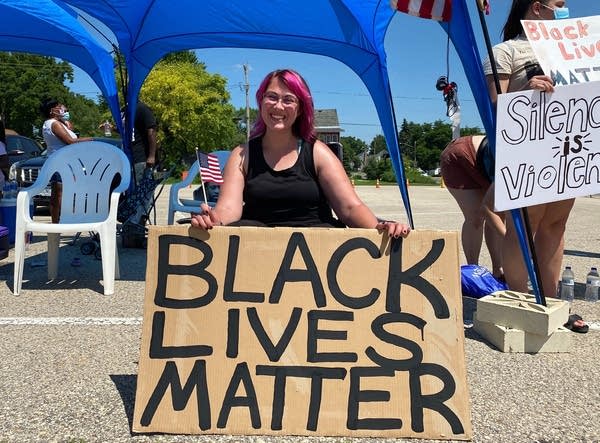 Person sits in a parking lot holding a sign that says "Black Lives Matter"