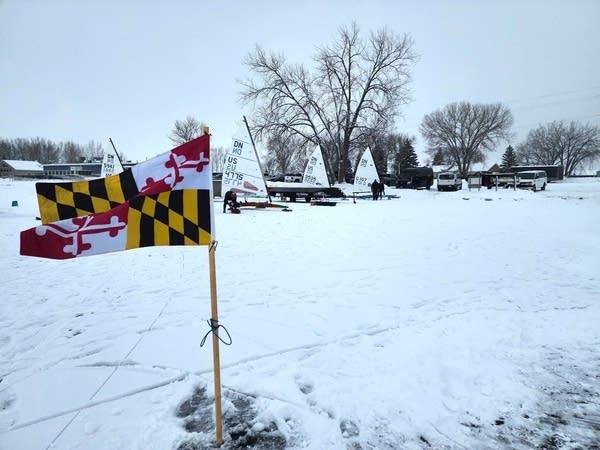 A flag flies in the wind while boats sit in the background with sails on top of them.