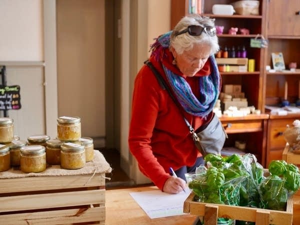 An older woman in a red shirt and blue scarf writes on a sheet of paper on a table, near containers of basil and honey.