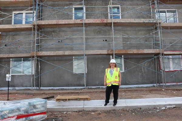A woman in a safety vest and hat stands in front of an apartment under construction