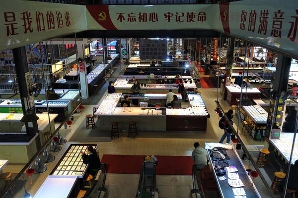 In a view from a high elevation, Ruili's Jiegao jade market is about half-way empty. There are roughly a dozen vendors sitting at tables with jade bracelets and other jade objects in front of them. A handful of customers, around three or four, are seen milling about.
