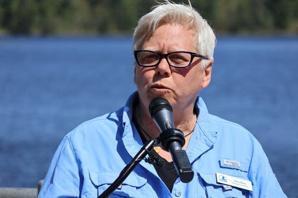 A man speaking at a podium with a body of water and trees in the background