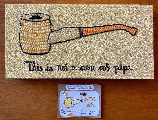 Overhead photo of crop art on a wooden table. It pictures a pipe made of seeds and the text, "This is not a corn cob pipe."
