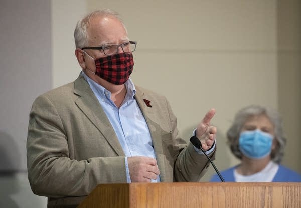 A man wearing a red and black plaid face mask gestures at a podium. 