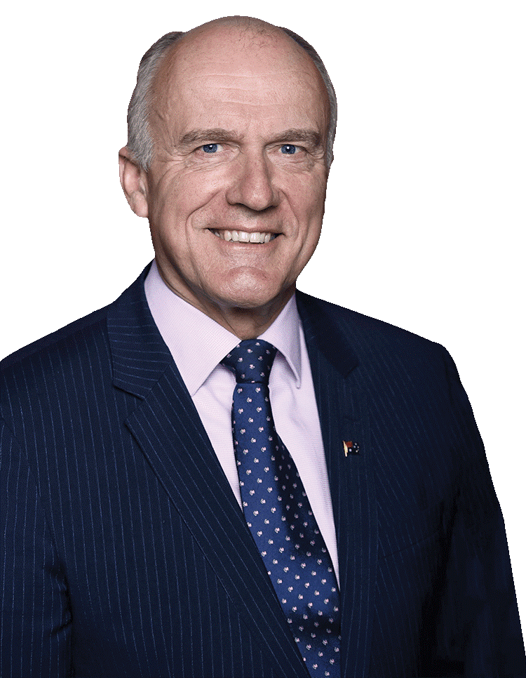 Eric Abetz, chairman of the Australian Monarchist League, is an older man sitting in front of a white backdrop. He is smiling and wearing a navy pinstripe suit, light pink button-up and blue tie.
