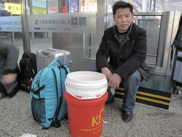 A man sits in the railway station with a few stacked plastic buckets and a blue backpack in front of him.