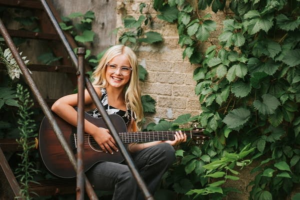 A musician sits with a guitar on a stairway near a building covered in vines.