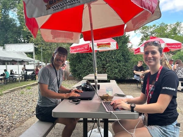 A man and woman smile for a photo while working on their laptops at a state fair table.