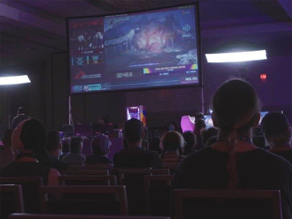 A crowd looks on to watch a video game speedrun projected on a large screen.