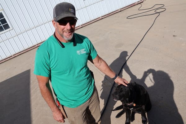 A man pets a black lab dog sitting nearby, while looking up at a camera.