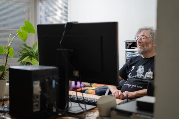 A man with gray hair sits at a desk behind a large desktop computer