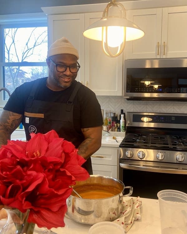 A man smiles in a kitchen next to a pot of orange soup.