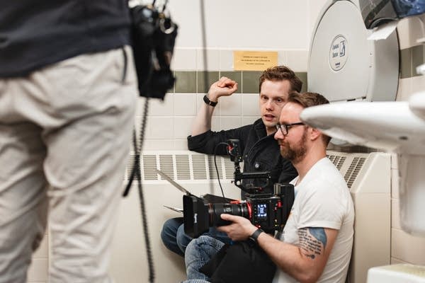 Two people talk while working on a filming session, kneeling inside a bathroom.