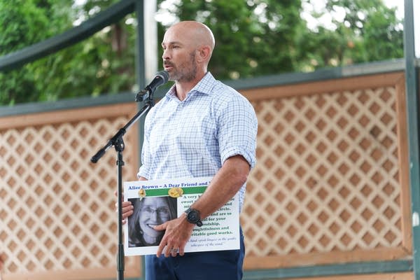 A man speaks into a microphone on a stage while holding a poster.