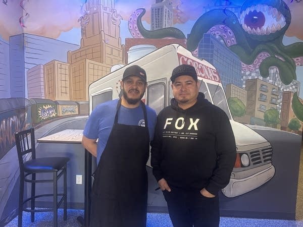 Two men pose next to a mural inside of a restaurant.