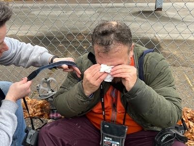 A man sits down on the sidewalk as others assist him.