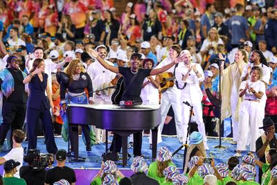 A musician at a grand piano spreads his arms on stage while flanked by other performers.