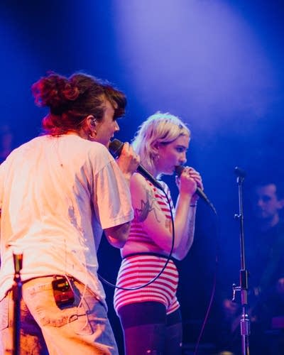 Two women sing into microphones on a lighted stage.