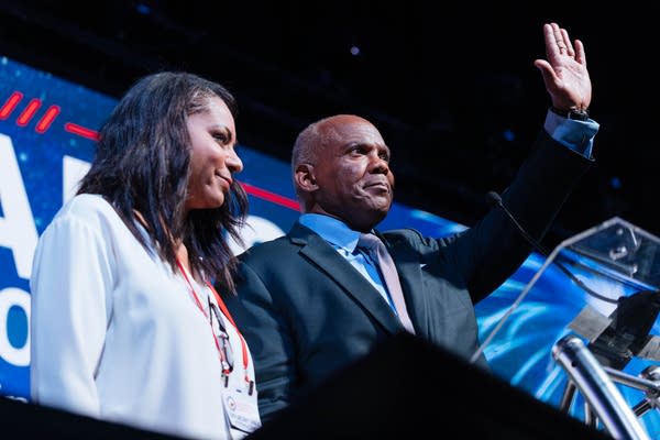A man raises a hand while on stage with a woman standing next to him.