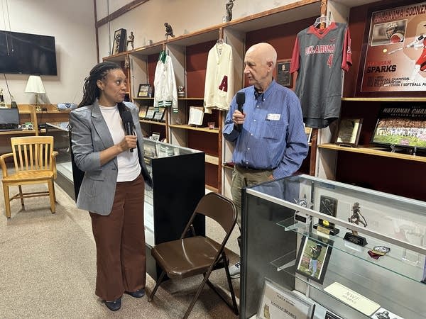 Marketplace Morning Report host Kimberly Adams stands with Charlie Anderson at the Thomasville Sports Hall of Fame. There are jerseys, trophies and other memorabilia on the shelves in the background.