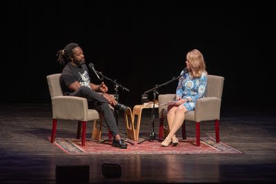 Two people sit on a stage with microphones.