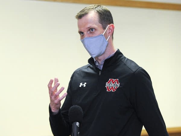 A man in a face mask talks in front of a microphone.