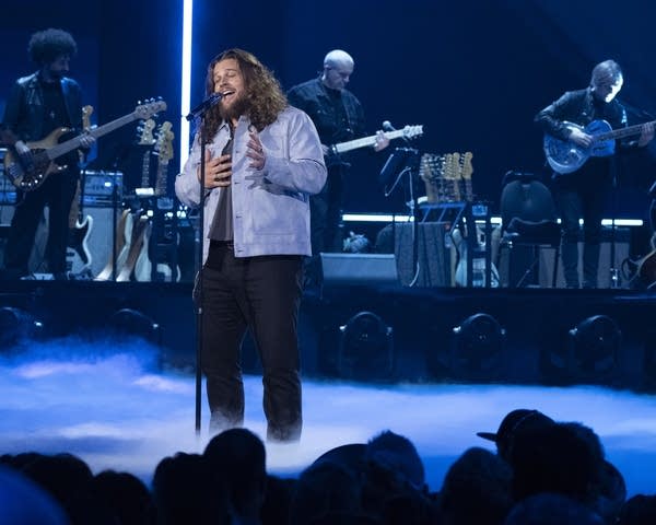 A man with long, curly hair sings on a blue-lit stage in front of a studio audience.
