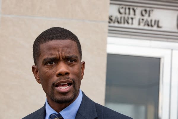 A man speaks into a microphone in front of a beige building. 