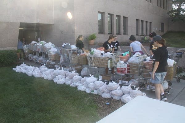 A row of shopping carts filled with food files out onto a sidewalk while stacked bags of food sit on the ground in a row.