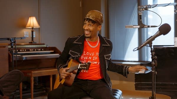A man smiles while playing piano with one hand and holding a guitar with the other