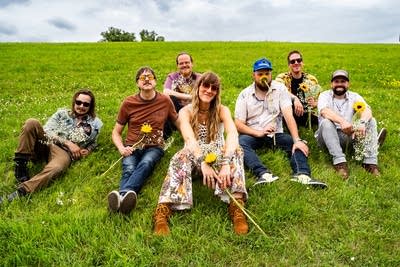A group of musicians, all holding sunflowers, sit together on a grassy hillside 