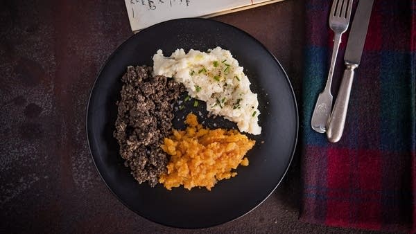 A black plate with three types of food. A tartan napkin with utensils on it sits to the right.