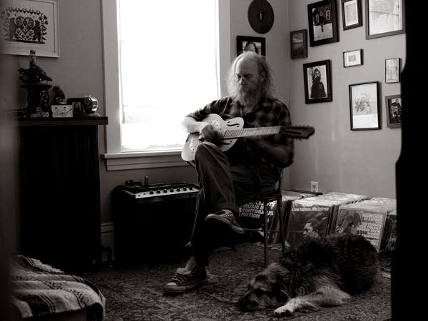 A man sits and plays a resonator guitar while a dog relaxes at his feet