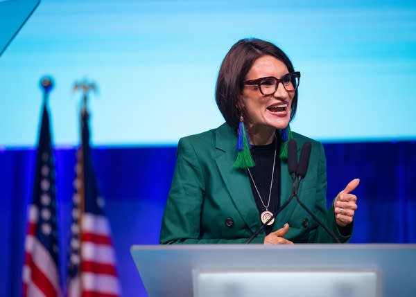 A woman in a green suit smiles and raises a thumb while speaking at a podium on stage.