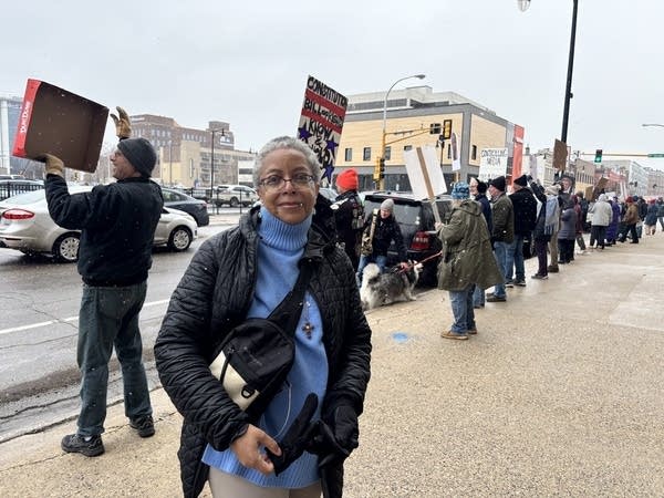 A woman poses for a photo in front of a group of protesters lined up on the sidewalk.