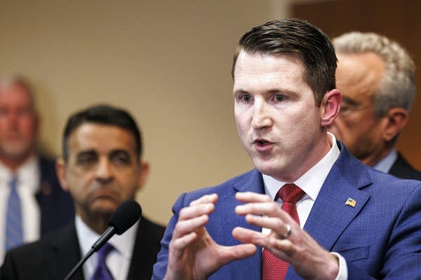 A man in a navy suit speaks at a podium during a press conference.