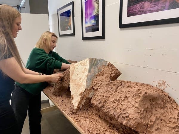 Two women work on a red clay base for a long sculpture.
