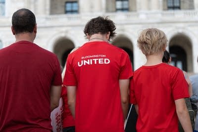 A man and two boys wear red shirts and have their backs to the camera. The middle shirt reads "Bloomington United."