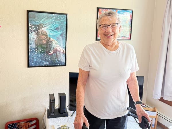 A woman using a walker smiles for a photo in her home. A framed poster of Jesus hangs on the wall.