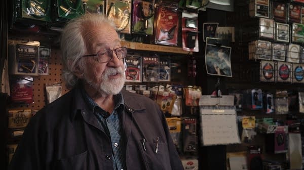 A bearded man behind a sales counter speaks to an interviewer