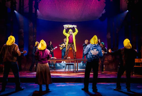 A man stands on a table on stage, surrounded by other performers.