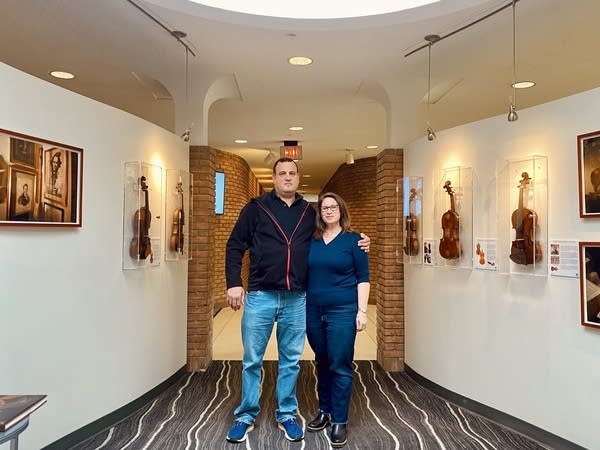Two people stand in a hallway with violins on display on the wall.