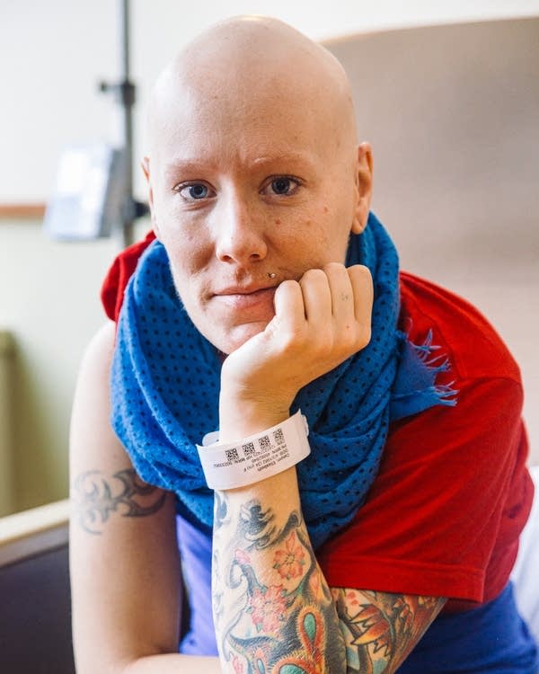 A woman with a shaved head, red shirt and blue scarf looks at the camera.