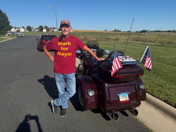 A man stands next to a motorcycle with a shirt that reads "Mark for Mayor."