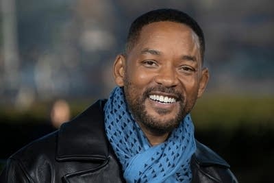 A man in a scarf and leather jacket smiles at the camera