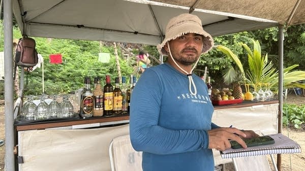 A man in a long-sleeved blue shirt and khaki hat stands in front of a beach shack bar.